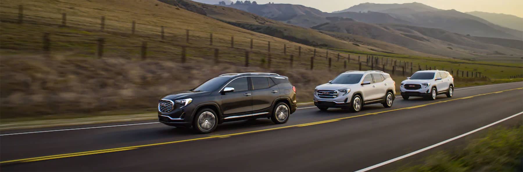 Three SUVs drive along a two-lane road in a rural area, with rolling hills and a fence in the background. The leading SUV is dark gray, followed by a silver one, and a white one trails behind. The setting is during the day.