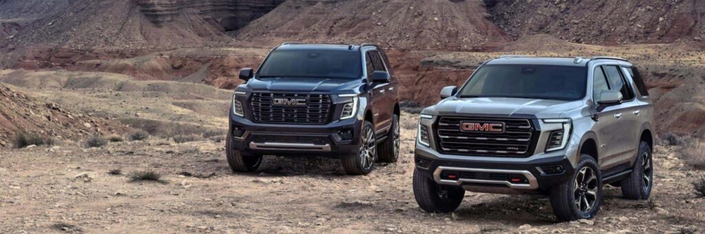 Two GMC SUVs parked on a rocky desert terrain with layered cliffs in the background. The vehicles are dark blue and silver, showcasing robust designs suitable for off-road adventures. The scene suggests a rugged, outdoor setting.