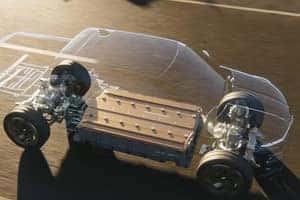 Transparent car chassis showing internal components, including the battery pack centrally located, attached to four visible wheels on an asphalt surface. The design emphasizes electric vehicle architecture.