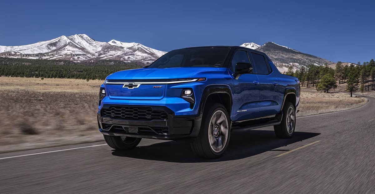 A blue pickup truck driving on a rural road with snow-capped mountains and a clear blue sky in the background. Trees are scattered along the horizon, and the road stretches into the distance.