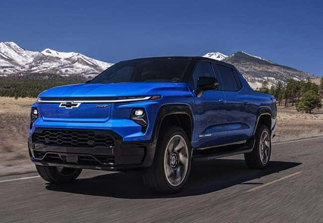 A blue Chevrolet pickup truck driving on a road with snow-capped mountains in the background under a clear blue sky.