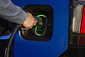 A person plugs a charging cable into an electric cars charging port, which is outlined with green lighting, against a blue vehicle body.
