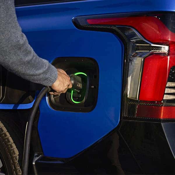 A person plugs a charging cable into the port of a blue electric vehicle. The rear light is visible, and the charging port is illuminated with a green light, indicating charging status.