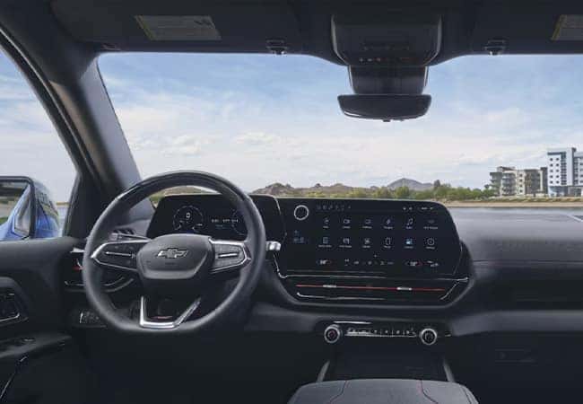 The image shows the interior of a Chevrolet vehicle, focusing on the sleek dashboard and steering wheel. A large digital display dominates the center console. Through the windshield, a landscape with buildings and mountains is visible.
