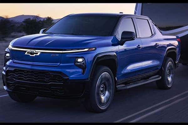 Blue Chevrolet pickup truck driving on a road at dusk, with blurred background indicating motion. The front grille features the Chevrolet emblem, and the truck is towing a trailer. The sky is transitioning from daylight to evening.