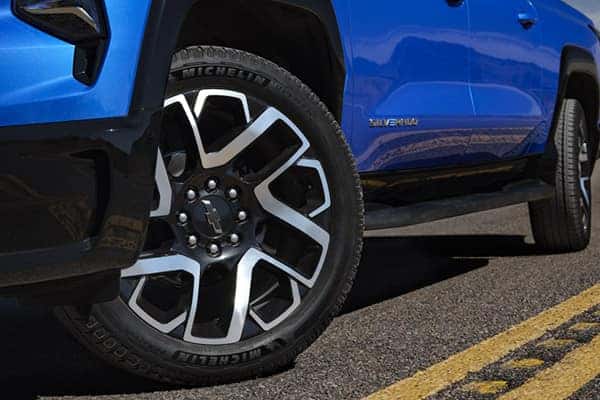Close-up of a blue vehicles front left wheel and tire on a road with yellow lines. The wheel has a distinctive multi-spoke design and the word Michelin is visible on the tire. The cars blue body is partly visible with a logo on the side.