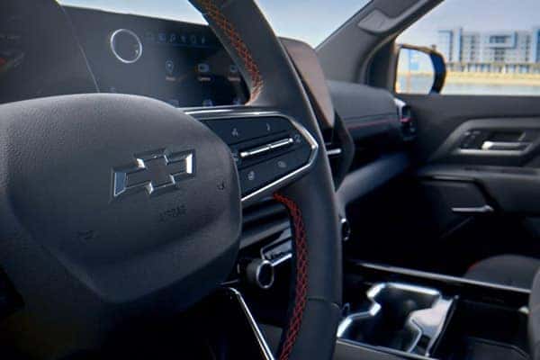 Close-up of a cars interior featuring a steering wheel with the Chevrolet logo. The dashboard has a digital display screen, and details of a modern infotainment system are visible. Sunlight illuminates the sleek design, and a building is in the background.