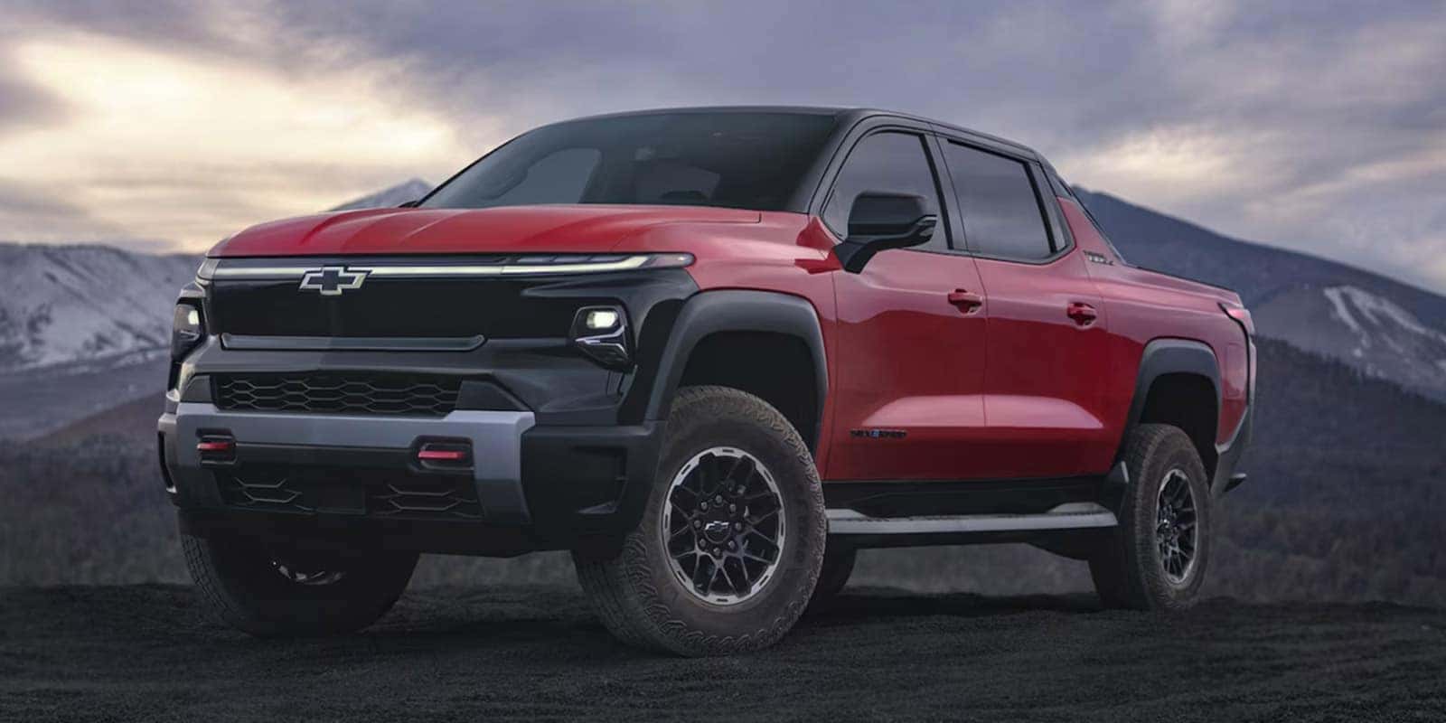 A red pickup truck parked on a dark, dirt surface with a mountainous backdrop. The sky is overcast, adding a dramatic effect to the scene. The truck has a rugged design with large tires and a prominent front grille.