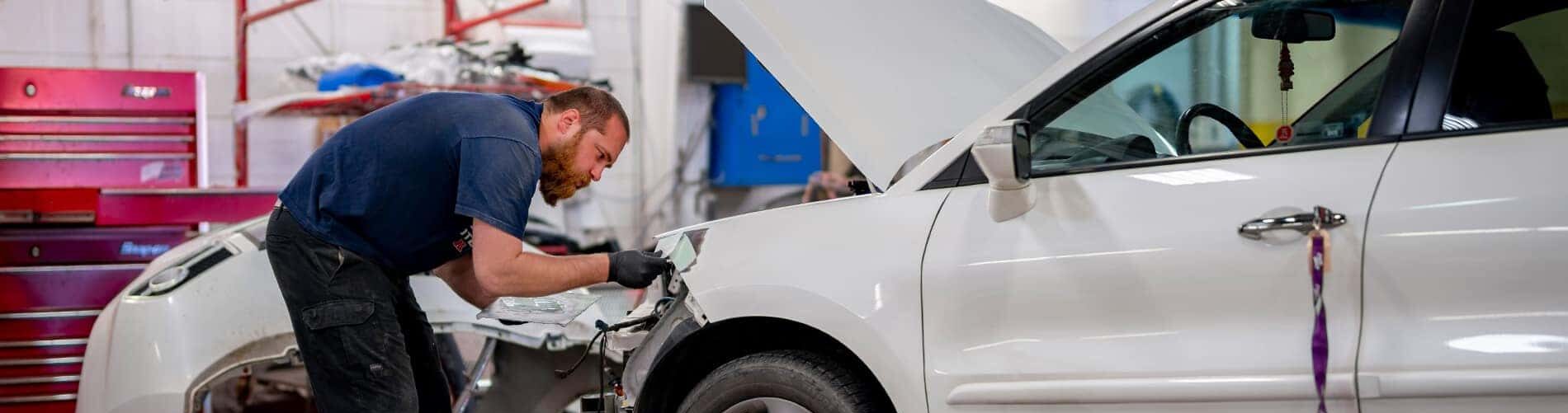 Mechanic repairing white car fender in auto repair shop, tools in background.