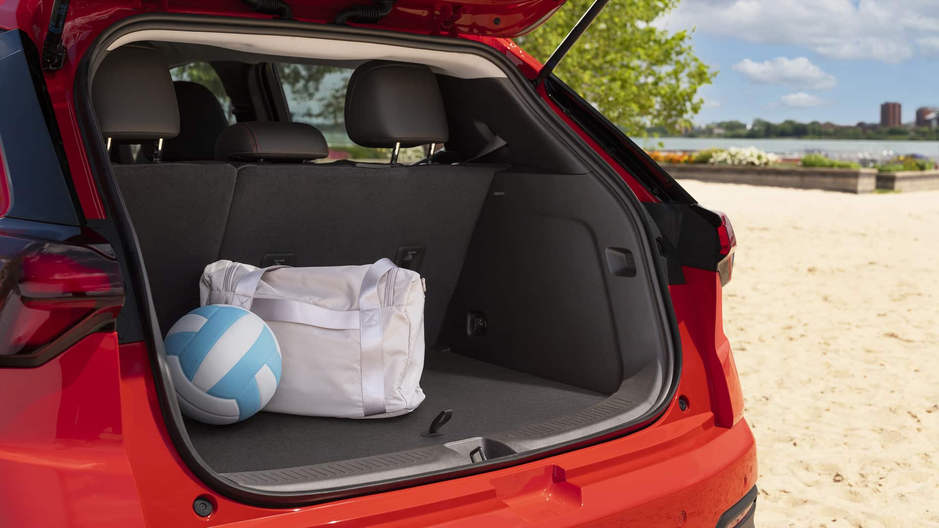 Open trunk of a red car with a white duffel bag and blue volleyball on sandy beach background.