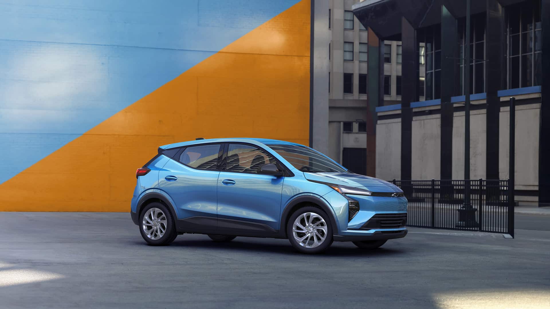 A red SUV is driving on an urban street. The vehicle features sleek, modern design elements with sharp headlights and a prominent grille. Buildings and trees are blurred in the background, suggesting motion.