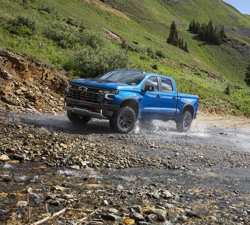 Blue pickup truck driving through a rocky stream in a lush mountain landscape.