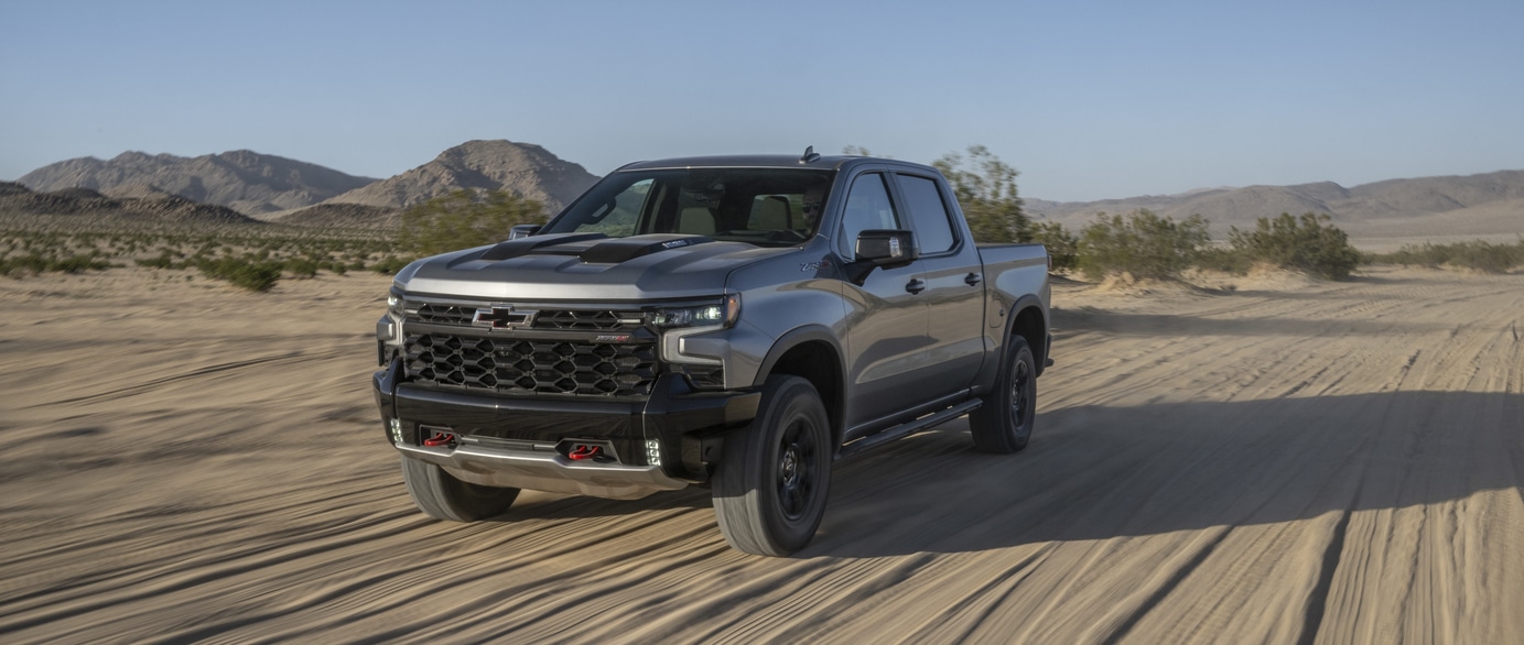 A green SUV is parked on a paved surface near a calm body of water. The car features a prominent grille and sleek design. In the background, hills are visible under a clear blue sky.
