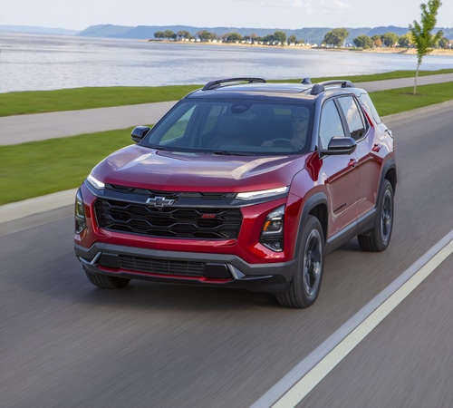 Red SUV driving on a scenic road by a lake, with trees and mountains in the background.
