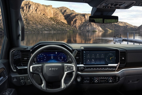 View from the backseat of a car showing two people in the front seats. The driver is holding the steering wheel. The sunroof is open, revealing a blue sky and tall buildings outside. The dashboard and rearview mirror are visible.