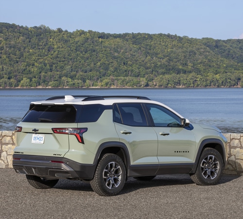 Green SUV parked by a lake with a forested hill in the background under a clear sky.