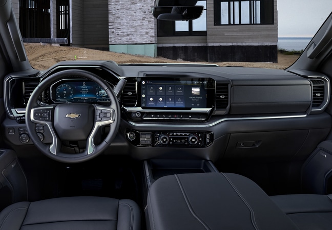 View from inside a car showing a cyclist crossing the street at an intersection in a city. The dashboard displays controls and information. Sunlight and shadows create a clear, bright scene.