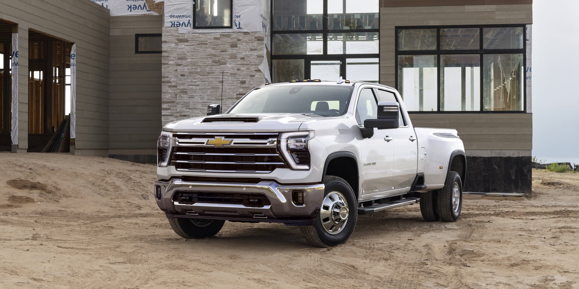 A black pickup truck drives through a sandy desert area, kicking up dust. The landscape features large rocks and Joshua trees under a clear blue sky. The truck has rugged off-road tires and a prominent grille.