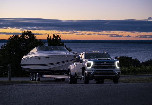 Truck towing a boat at sunset by a lake, with silhouetted trees and a vibrant sky.