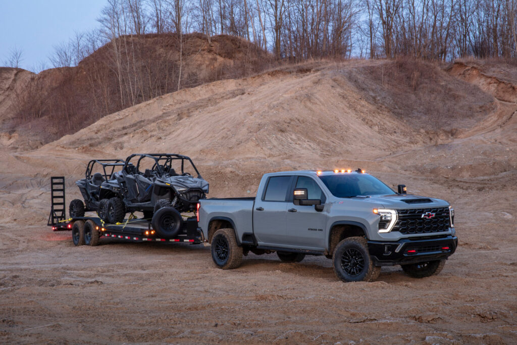 A gray pickup truck towing two off-road vehicles on a trailer in a sandy, hilly landscape.