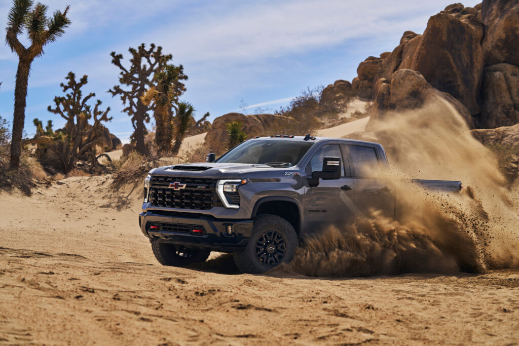 Heavy-duty pickup truck driving fast through desert sand, surrounded by rocky terrain and sparse vegetation.