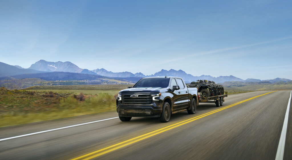 Black pickup truck towing ATVs on a highway with scenic mountains in the background.