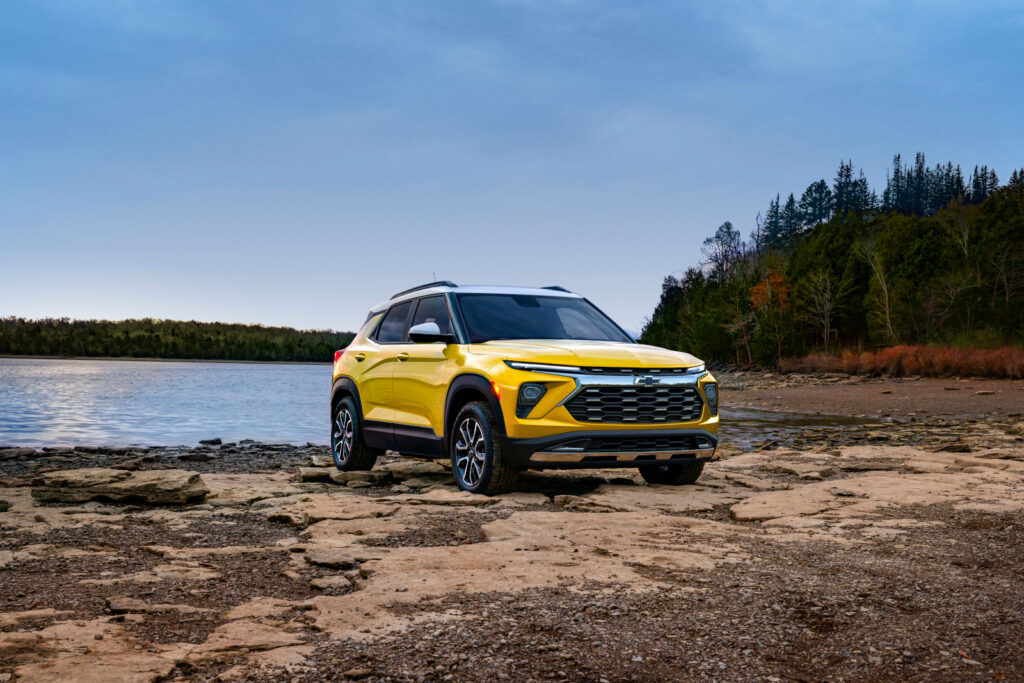 Yellow SUV parked by a serene lakeshore, surrounded by forested landscape under a blue sky.