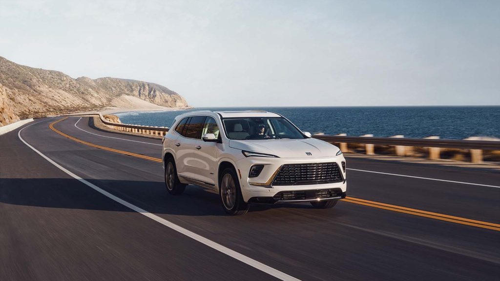 White SUV driving on a coastal highway with ocean and cliffs in the background.
