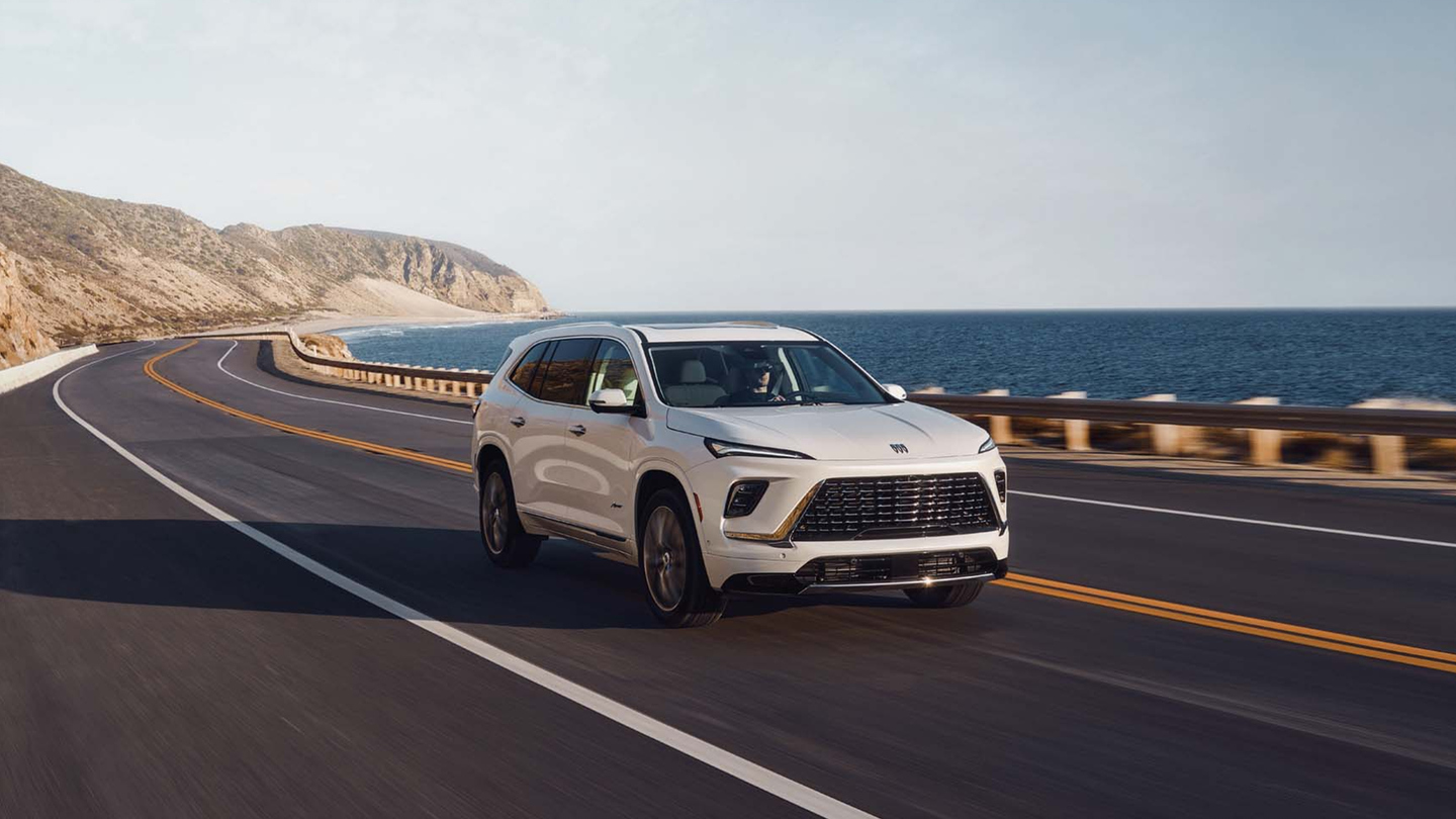 White SUV driving on a coastal highway with ocean and cliffs in the background.