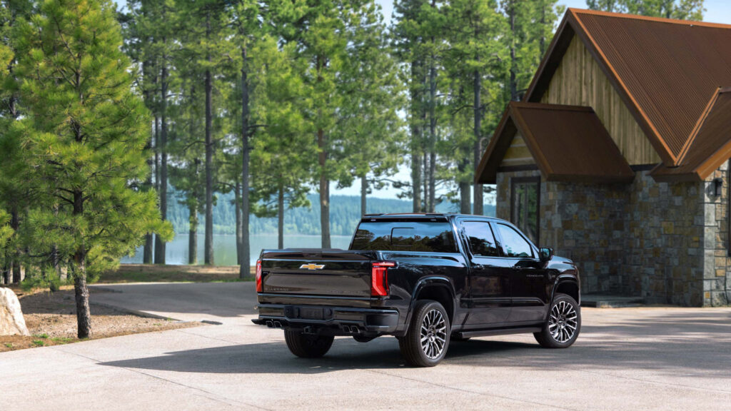 Black pickup truck parked on a driveway near a house surrounded by tall trees and a lakeside view.